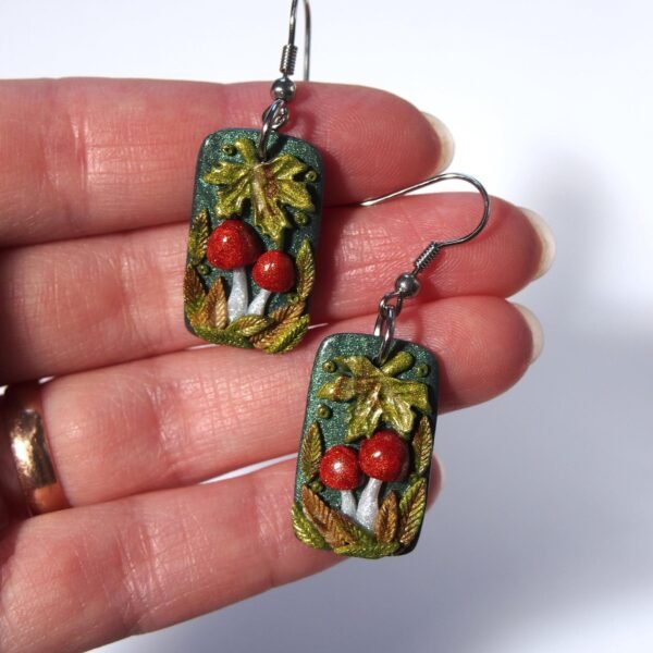 A pair of handmade polymer clay mushroom earrings. The rectangular dangle earrings feature two detailed red mushrooms, nestled among shiny, multi-tonal autumn leaves on a dark green base. This one-of-a-kind (OOAK) piece of whimsical cottagecore jewelry has a distinct fairycore and woodland aesthetic. The earrings are finished with surgical steel hooks, making them a unique artistic gift for a nature lover.