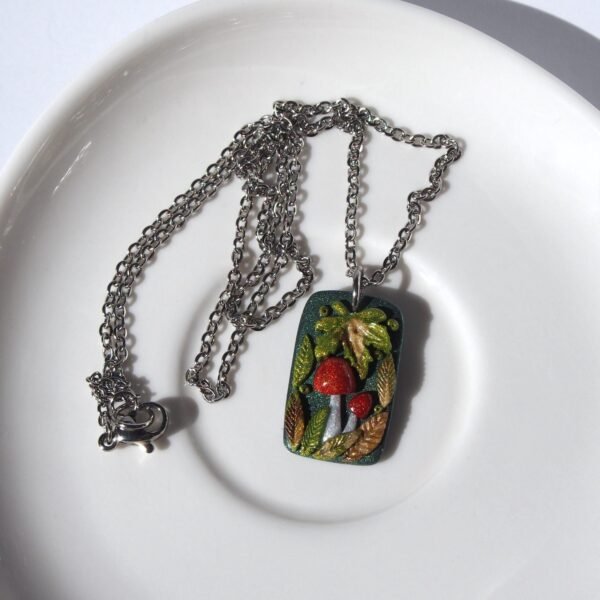A handmade polymer clay mushroom pendant necklace. The rectangular design features two vibrant red toadstools nestled among shiny, multi-tonal autumn leaves on a dark green textured base. This one-of-a-kind (OOAK) piece of whimsical cottagecore jewelry has a strong fairycore and woodland aesthetic. The pendant hangs from a stainless steel chain, making it a unique, artistic nature-themed gift.