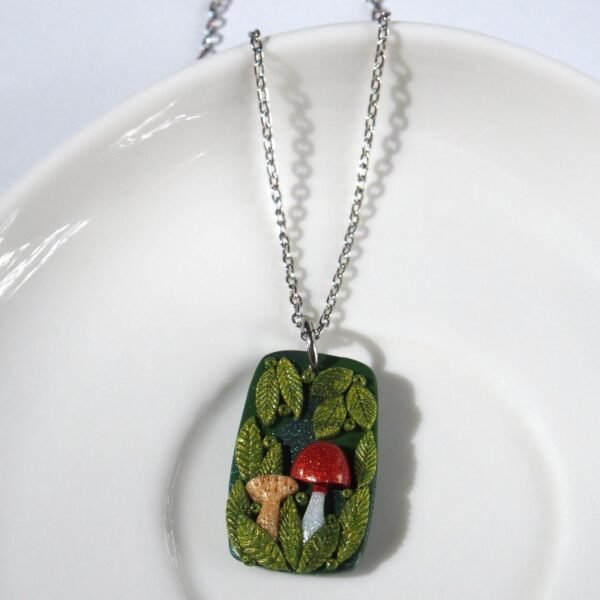 A handmade polymer clay mushroom pendant necklace. The rectangular design features a charming forest scene with a red toadstool, a beige mushroom, and lush, shiny green leaves on a dark, sparkling base. This one-of-a-kind (OOAK) piece of whimsical cottagecore jewelry has a strong fairycore and woodland aesthetic. The pendant hangs from a stainless steel chain, making it a unique, artistic nature-themed gift for her.