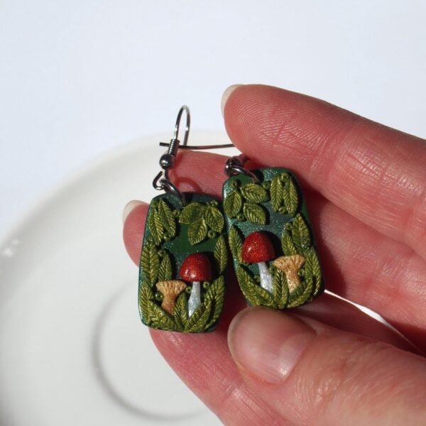A pair of handmade polymer clay mushroom earrings. The rectangular dangle design features a charming forest scene with a red toadstool, a beige mushroom, and lush, shiny green leaves on a dark, sparkling base. This one-of-a-kind (OOAK) piece of whimsical cottagecore jewelry has a strong fairycore and woodland aesthetic. They are finished with hypoallergenic surgical stainless steel hooks, making them a unique, artistic nature-themed gift.